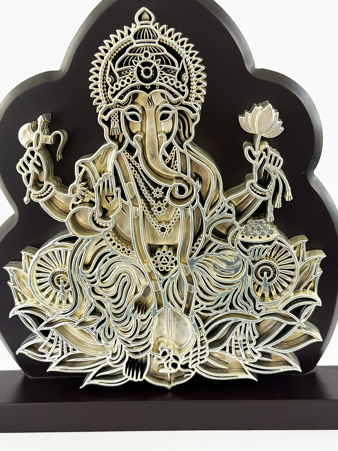 Intricate metal relief of Hindu god Ganesha seated on a lotus with a detailed crown and multiple arms holding symbolic items on a dark plaque.