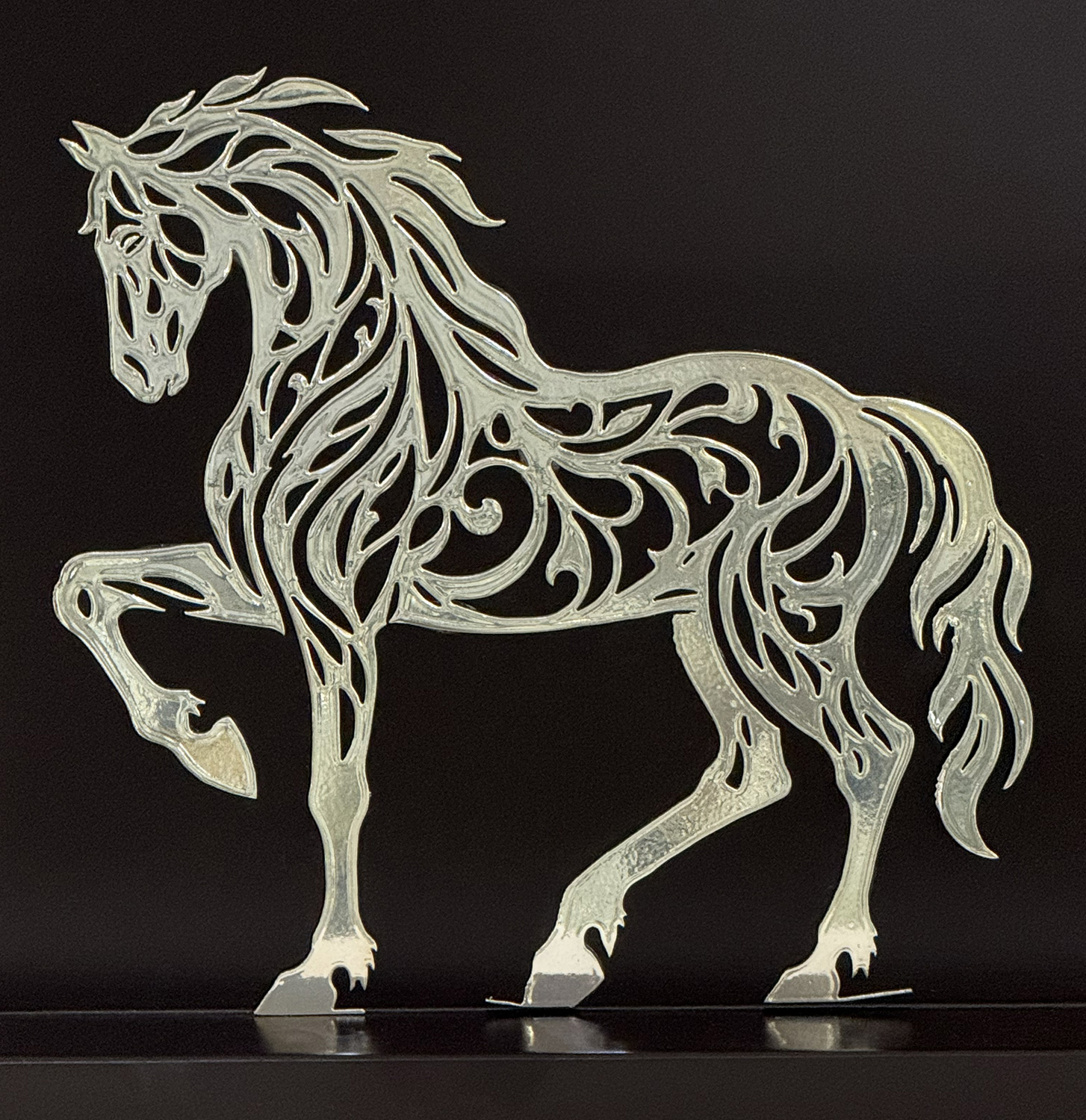 Ornate metal horse silhouette with swirling filigree patterns against a dark background, decorative art piece.