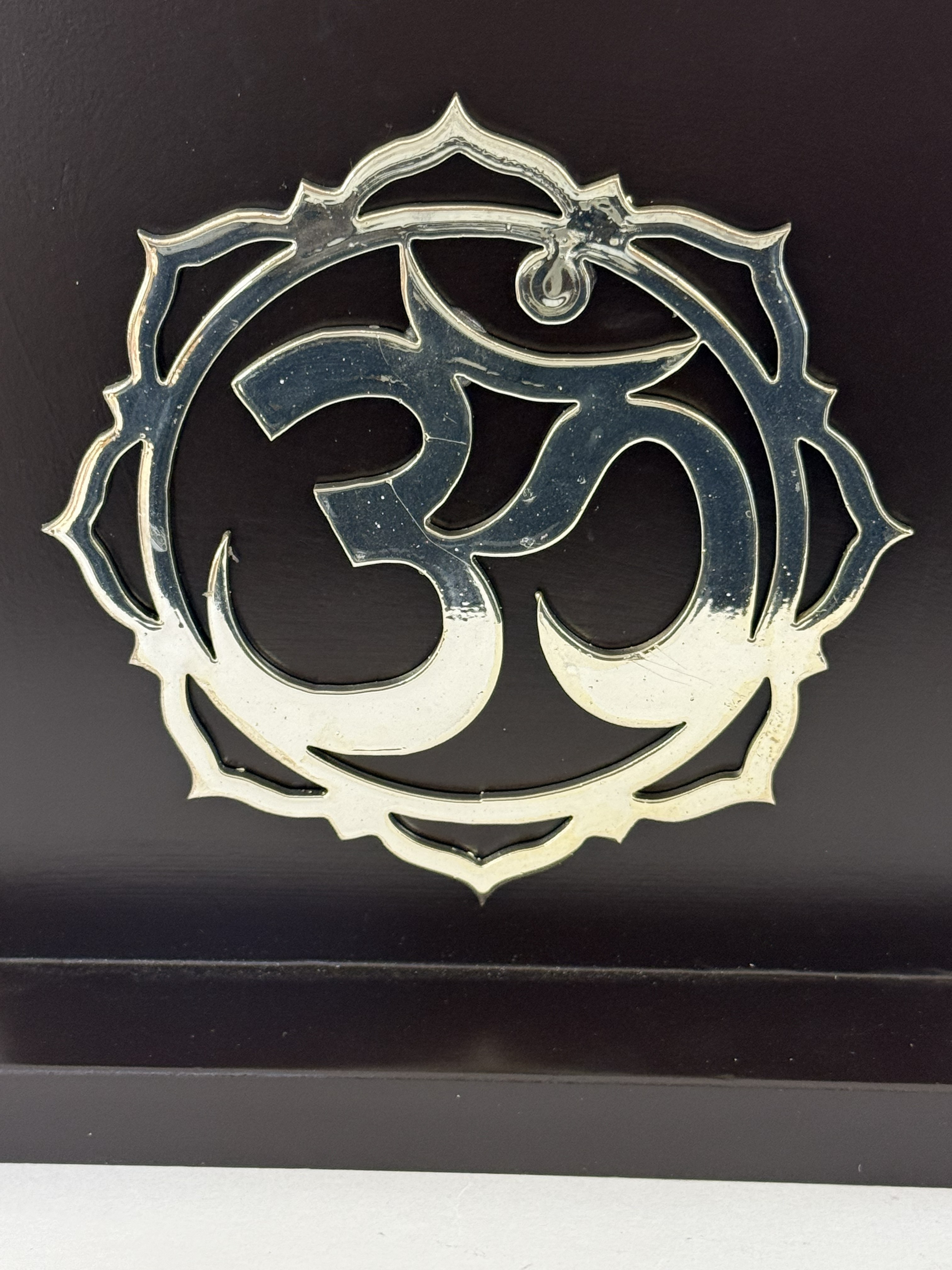 Decorative metal Om symbol within an ornate circular frame on a dark surface (baroque-inspired emblem).