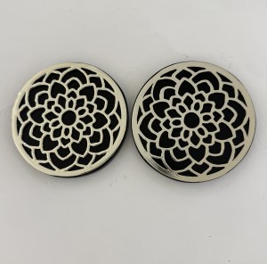 Two round metal coasters with an intricate black floral lattice pattern side by side in a pale metal frame.