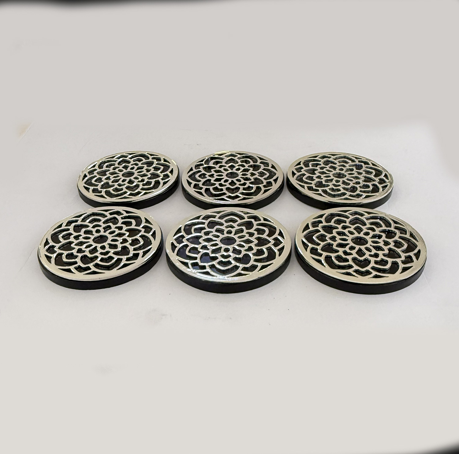 Six round metal coasters with an intricate lace-like floral cut-out design, arranged on a light surface.