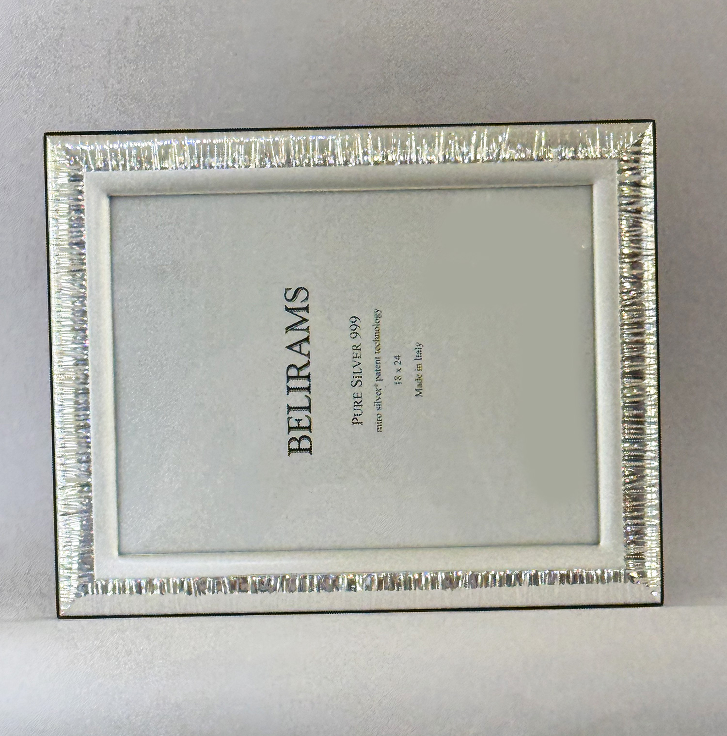 Close-up of a decorative silver ribbed picture frame with a pale gray inner mat and the word BELIRAMS printed inside.