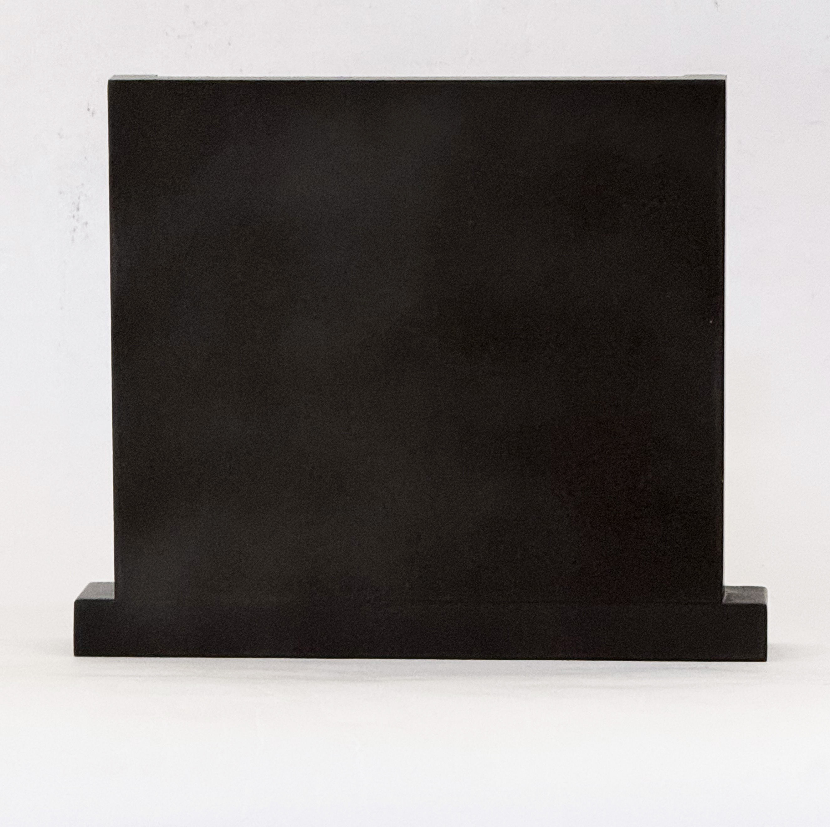 Large black rectangular block resting on a small base against a light wall, photographed head-on on a white surface.
